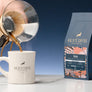 A 12oz bag of 700 beside a diner mug as coffee is being poured from a chemex into the mug, on a marble counter