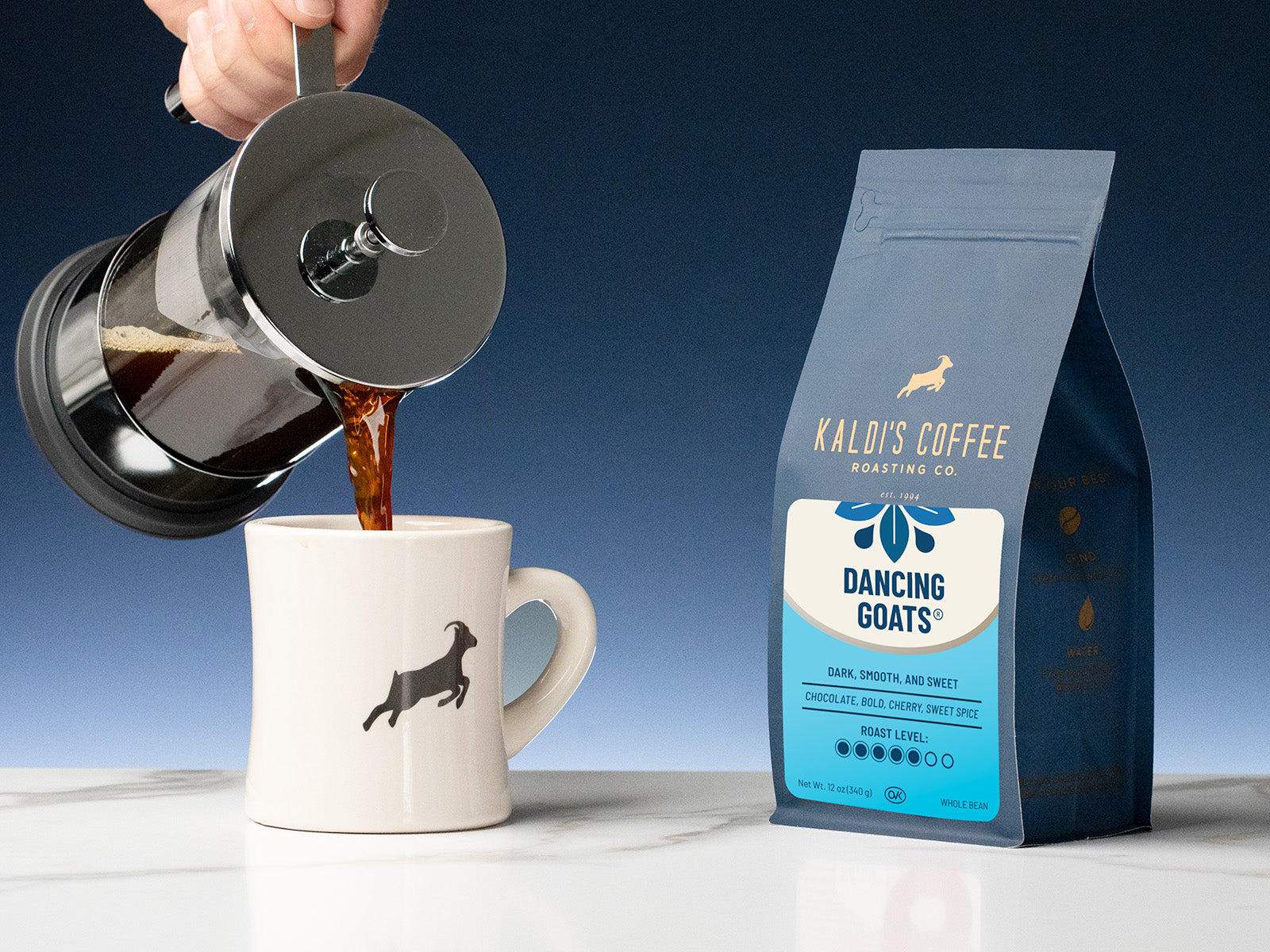 A french press pours coffee in a diner mug, sitting beside a 12oz bag of Dancing Goats Blend on a marble counter