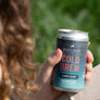 Close-up of a hand holding a chilled can of Kaldi's Coffee Cold Brew, Classic Black, with a blurred background of green grass. The can features pink "COLD BREW" lettering.