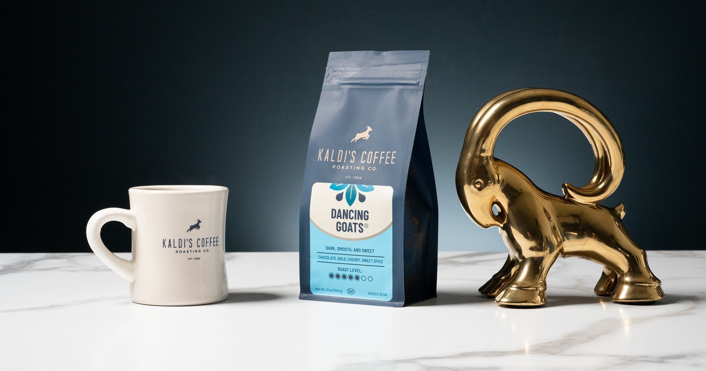 A small bag of Kaldi's Coffee "Dancing Goats" coffee, a white Kaldi's Coffee mug, and a gold goat figurine on a marble surface against a dark background.