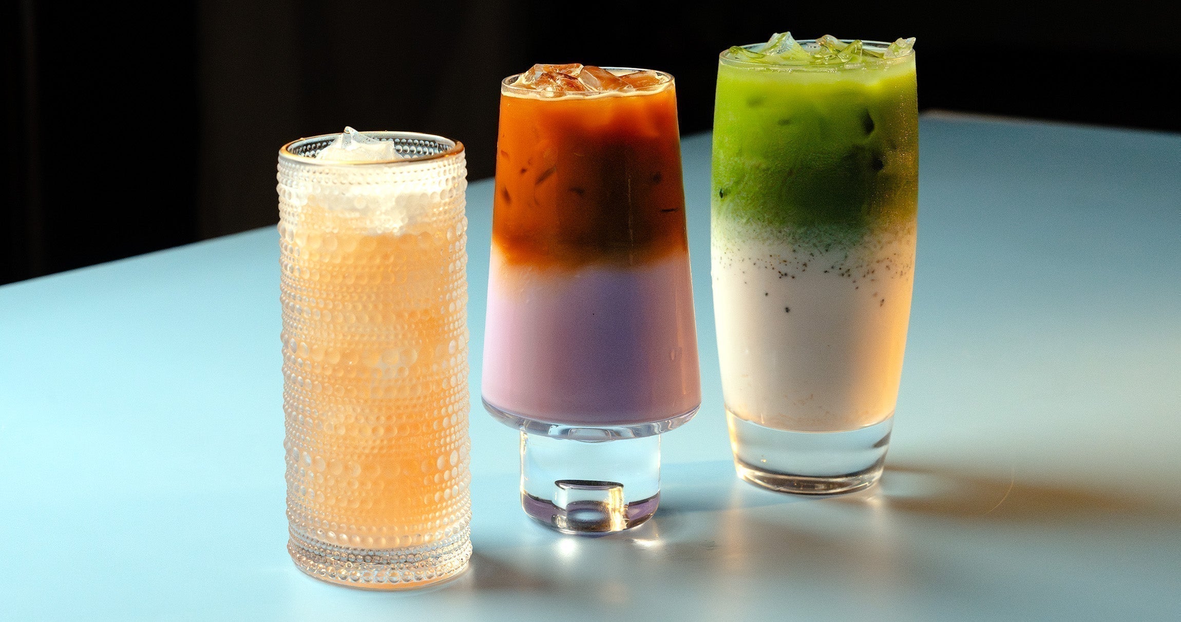 3 drinks sit side by side on a blue table. They are grapefruit colored, purple and brown, and green and white layered drinks, from left to right