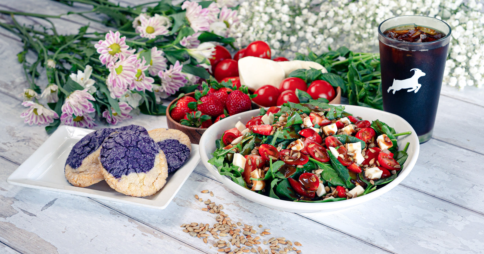 A table of beach wood is covered in flowers, basil, mozzarella, and tomatoes. In the midst of them, a plate of purple ube cookies, a Caprese salad, and a glass of cold brew sit, ready to be consumed