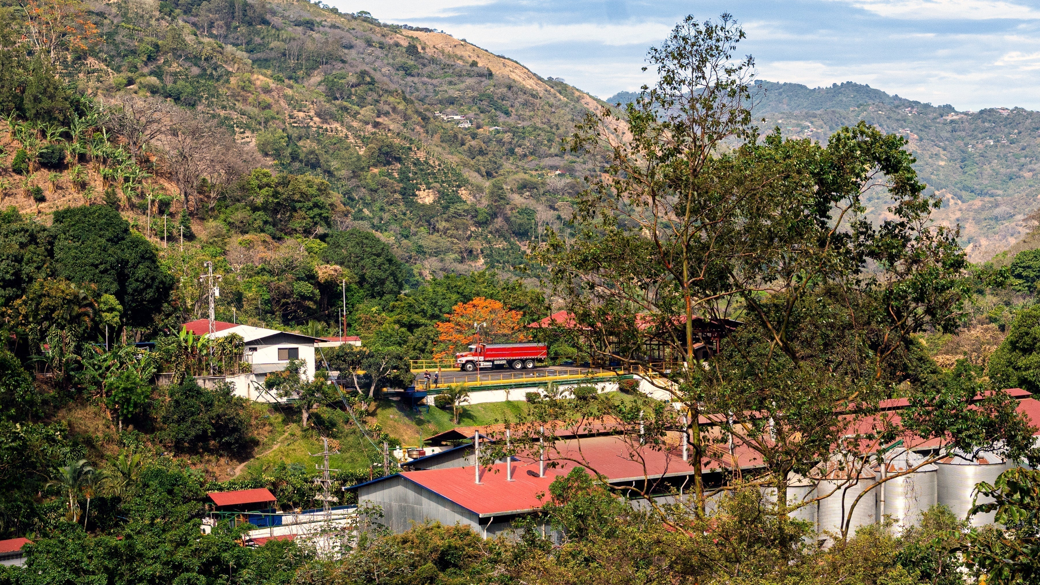 An industrial complex lies in a valley, silos, red roofs, and buildings visible. In the center, a large, red Mack truck is parked on a flat strech of pavement