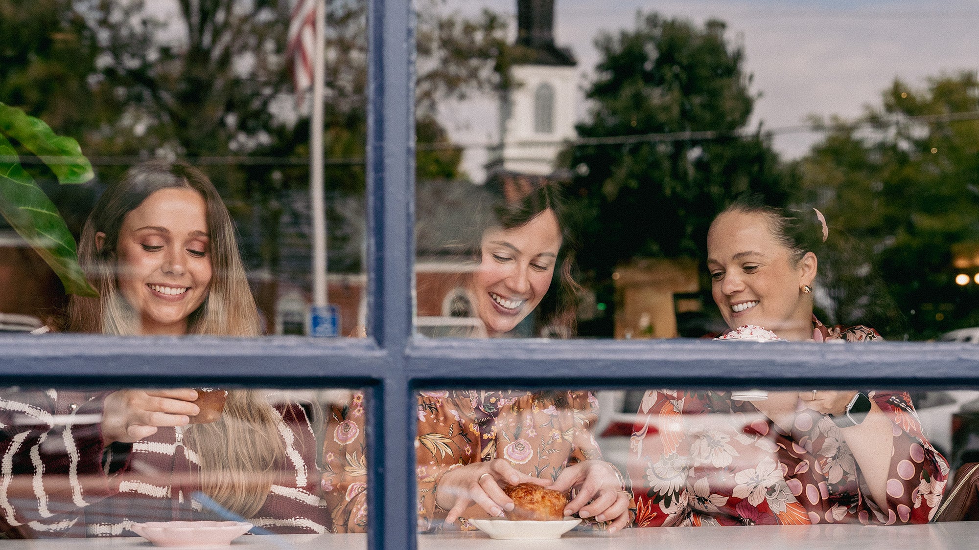 Three women sit at a counter, holding drinks and pastries as they look down and smile. They sit behind a window, a flag pole, trees and a spire visible in the reflection of the glass.
