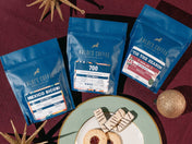 Three blue coffee bags from Kaldo's Coffee on a dark red surface with a plate of cookies.