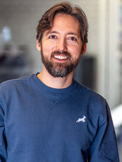 A smiling man with a beard wears a blue sweatshirt with Kaldi's goat logo over his left breast