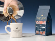 A 12oz bag of 700 beside a diner mug as coffee is being poured from a chemex into the mug, on a marble counter