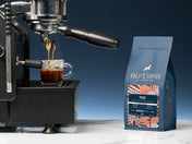A shot of espresso pours from an espresso machine in profile, on a white marble counter with a blue wall behind. Beside the espresso machine sits a 12oz bag of 700.