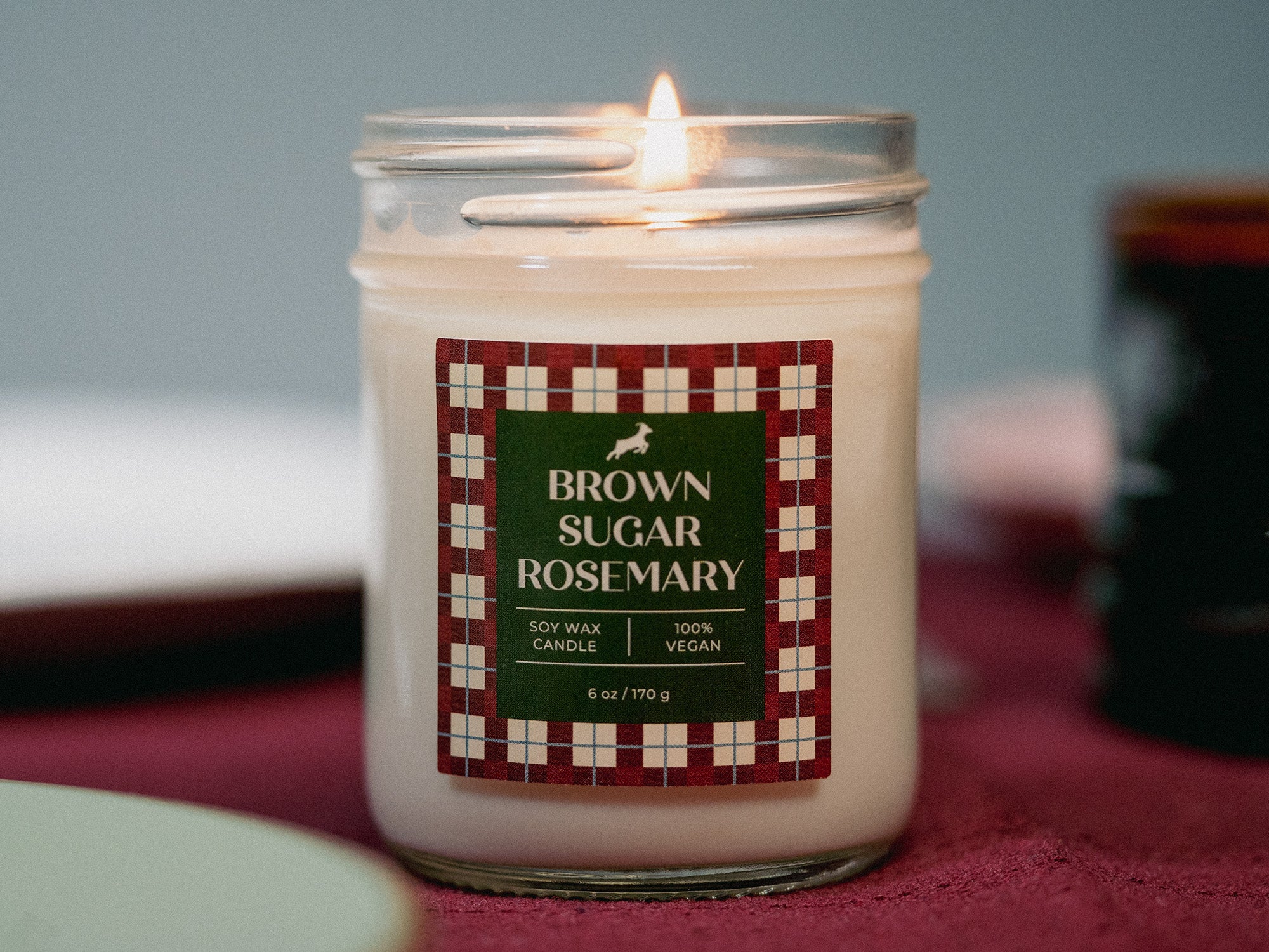 Candle labeled 'Brown Sugar Rosemary' in a glass jar on a red surface with a blurred background.