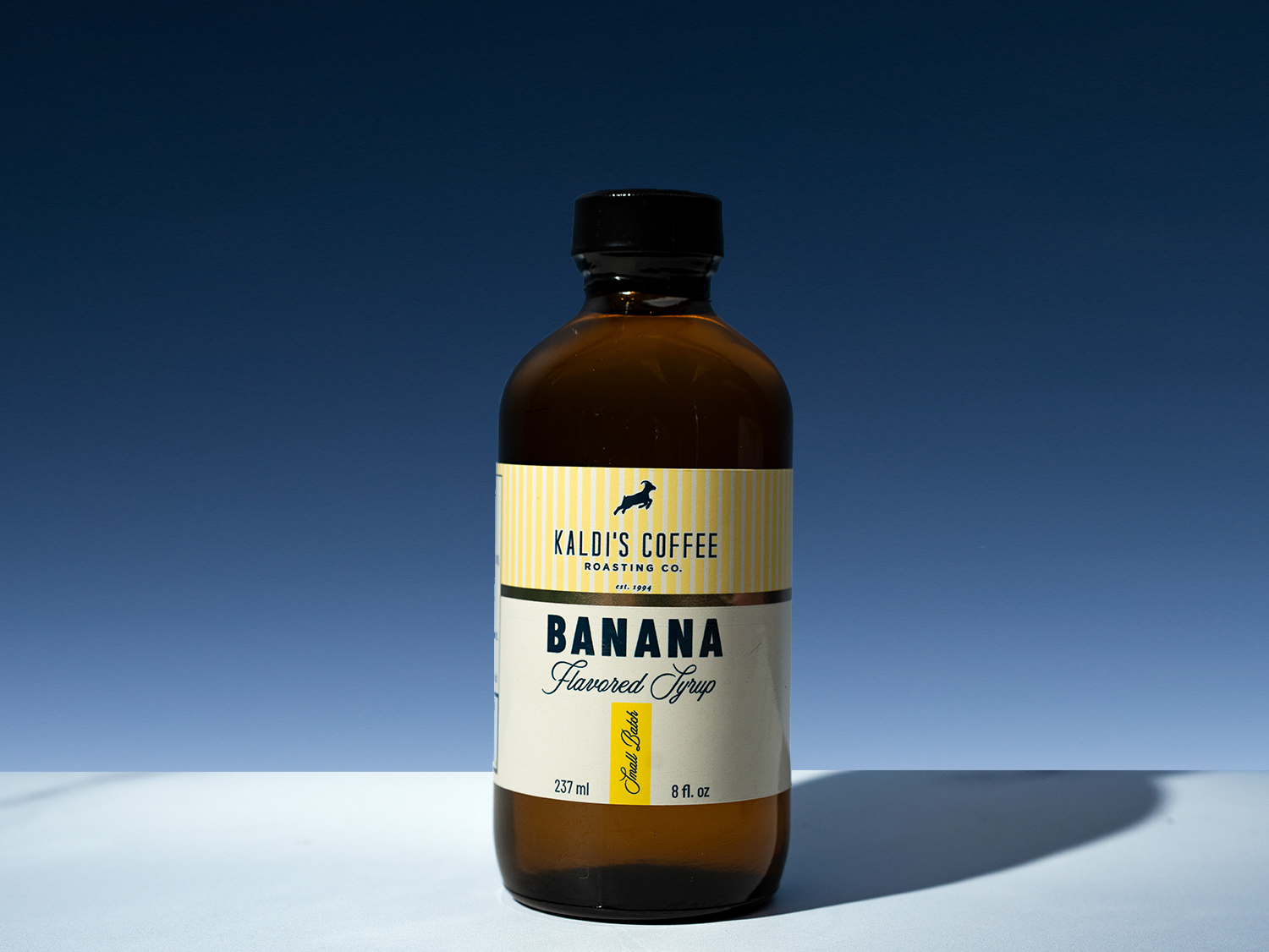 A bottle of Kaldi's Coffee Roasting Co. Banana Flavored Syrup, in an amber glass bottle with a black cap and a cream and yellow label, set against a dark blue gradient background.