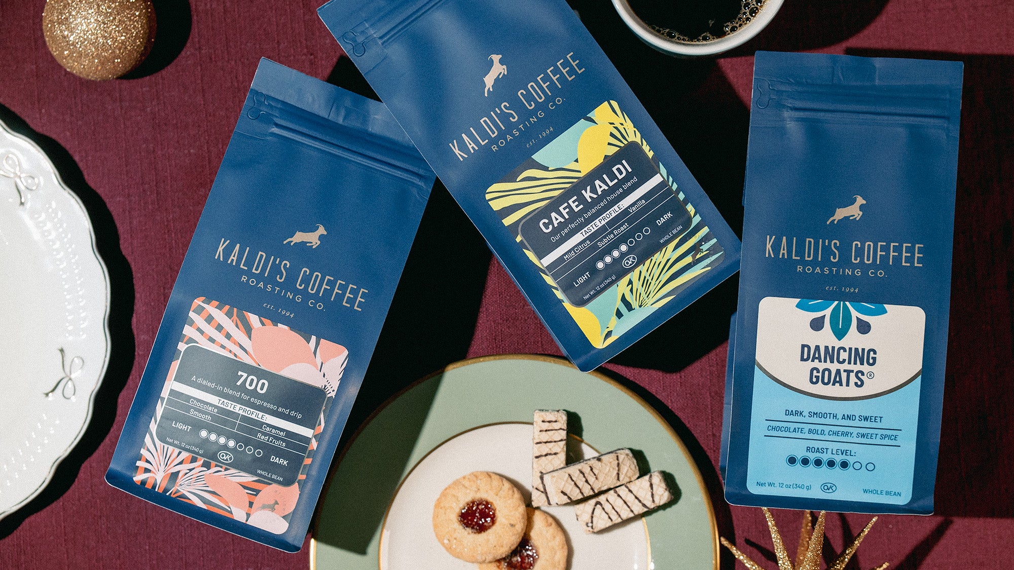 Three packages of Kaloi's Coffee on a dark surface with cookies and a cup of coffee.