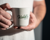 A person holding a white ceramic coffee mug with 'Kaldi's Coffee' printed on it.