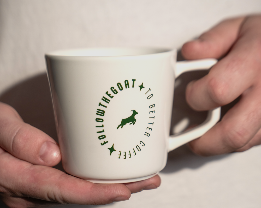A white ceramic coffee mug with a green circular graphic reading 'FOLLOW THE GOAT TO BETTER COFFEE'.