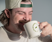 A man drinks from a white ceramic mug featuring a green circular logo with a goat silhouette and text.