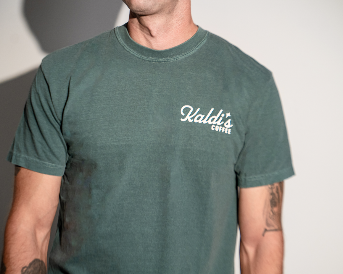 A man wearing a vintage dark green crewneck t-shirt with a white 'Kaldi's Coffee' logo.