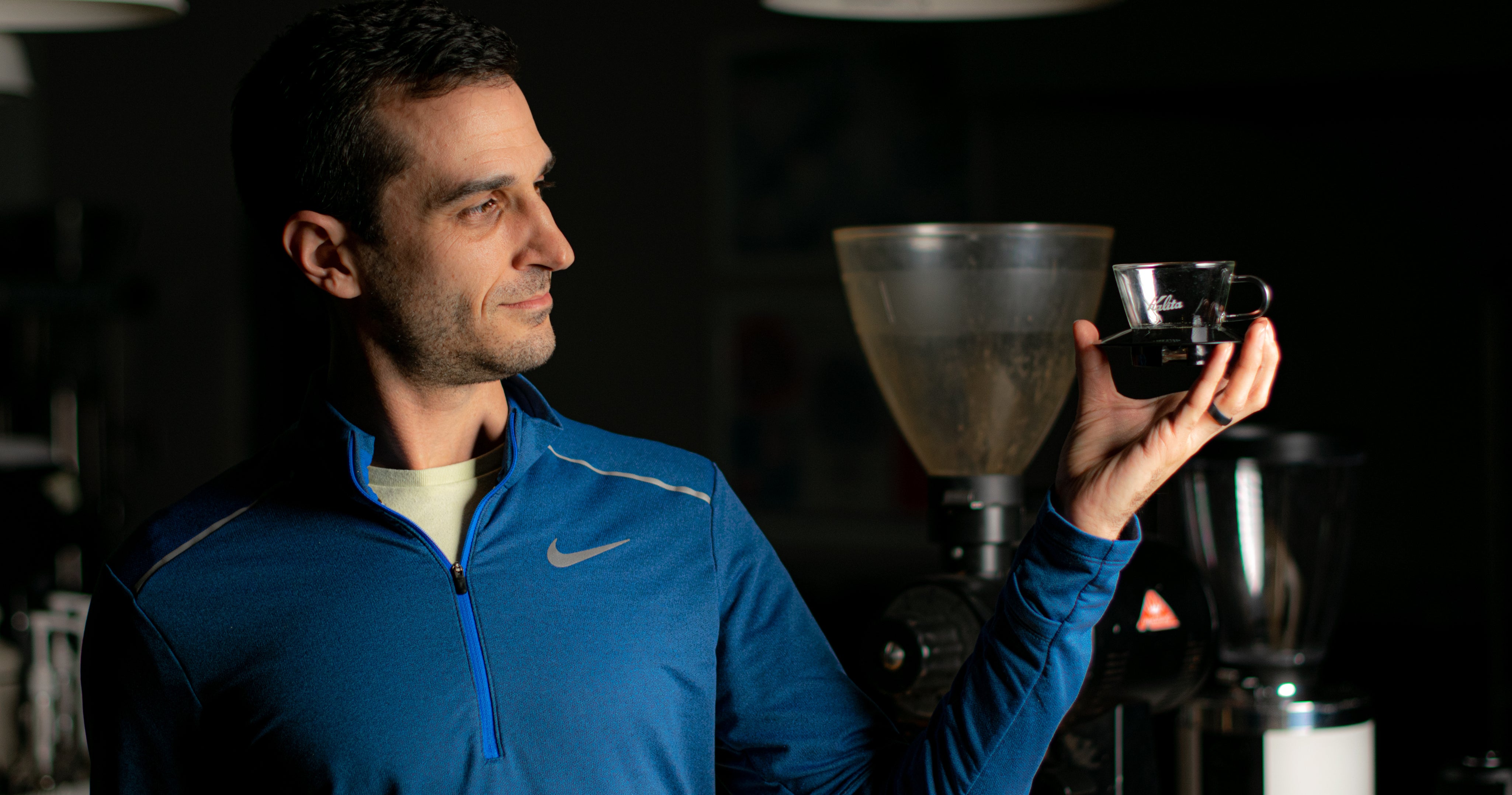 Man in blue jacket holding a coffee cup and funnel in a dark setting