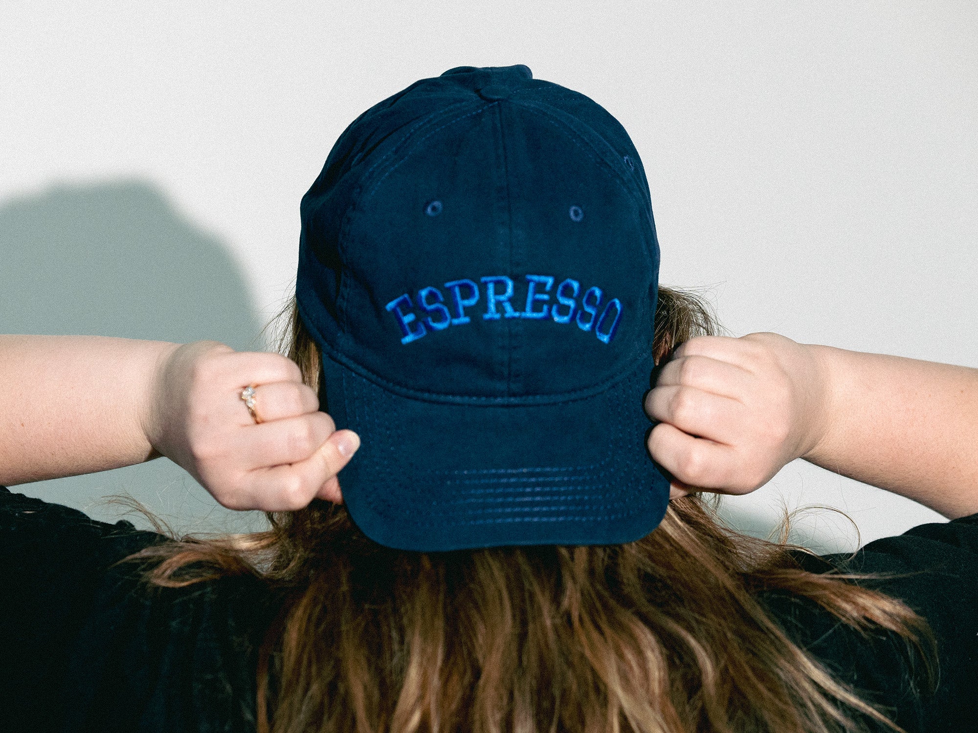 Person wearing a navy blue cap with 'ESPRESSO' text, holding it behind their head against a plain background.