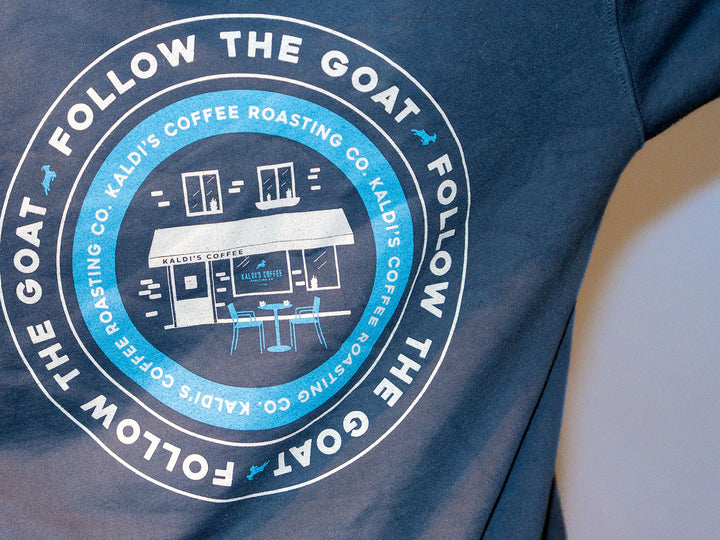 A close up of a blue sweatshirt's back, with a circular "follow the goat" imprint, surrounding an illustration of a cafe and the words "Kaldi's Roasting Co" repeated in the same circle