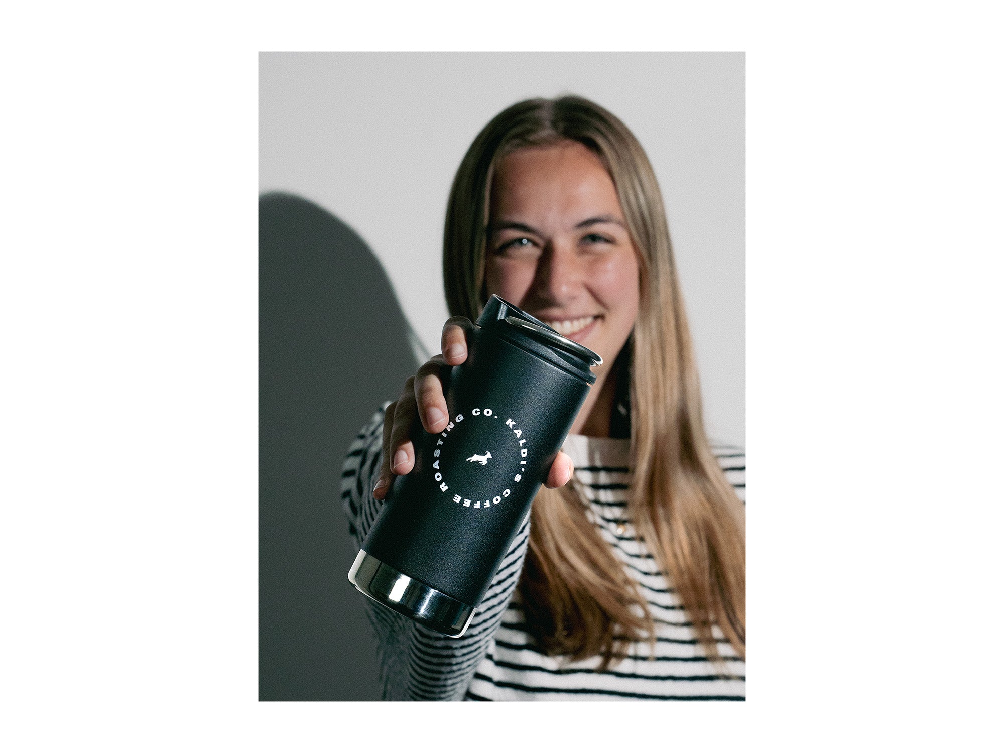 A smiling woman holds a black Klean Kanteen with Kaldi's logo, stretching her arm toward the camera