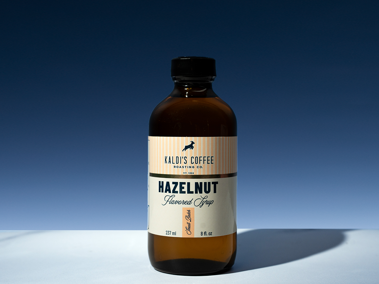 A bottle of Kaldi's Coffee Roasting Co. Hazelnut Flavored Syrup, 8 fl oz, in an amber glass bottle with a black cap and cream label, set against a blue gradient background.