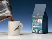 A coffee pot pours into a diner mug, sitting on a marble counter beside a 12oz bag of Highlander Grogg