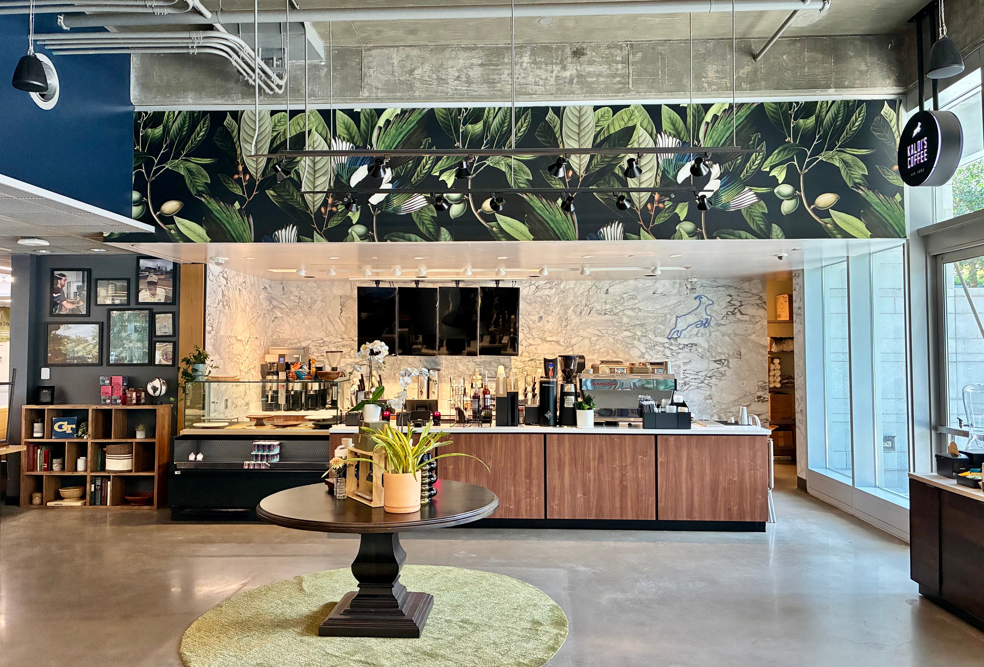A modern interior space with a large decorative wall mural featuring green leaves and black birds, above a kitchen or cafe counter with wood cabinets, coffee equipment, shelving, and a round table in the foreground.