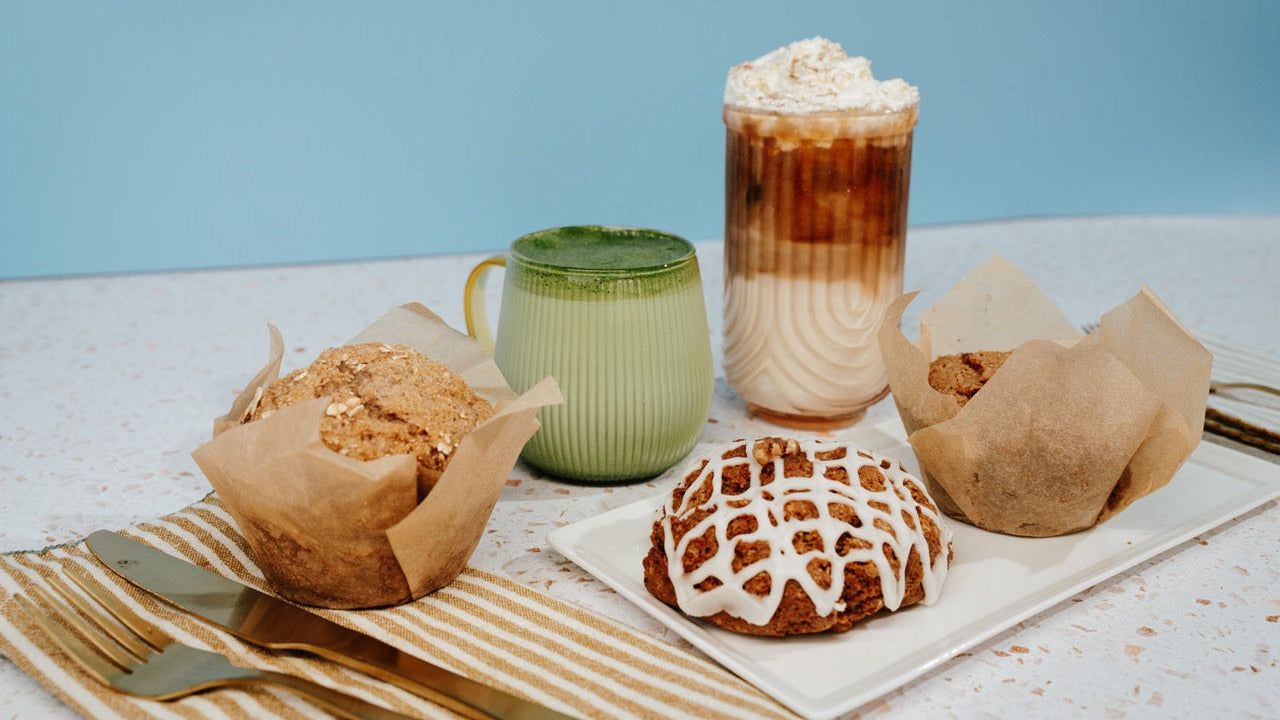 A styled food and beverage spread on a layered iced coffee drink with whipped cream, and three pastries in brown paper liners, including one with white icing drizzle.