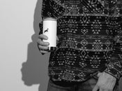 Person wearing a patterned shirt holding a white tumbler with a logo.