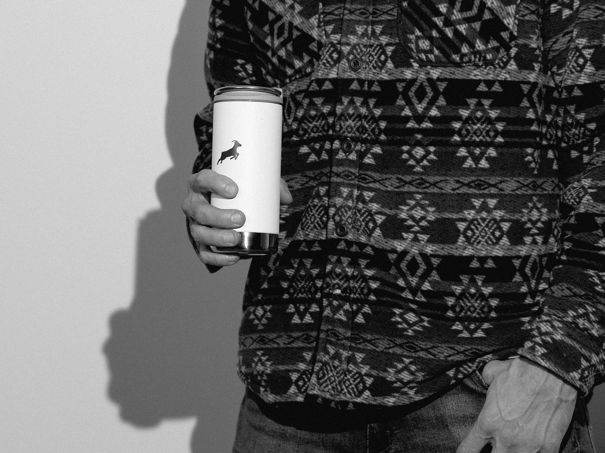 Person wearing a patterned shirt holding a white tumbler with a logo.