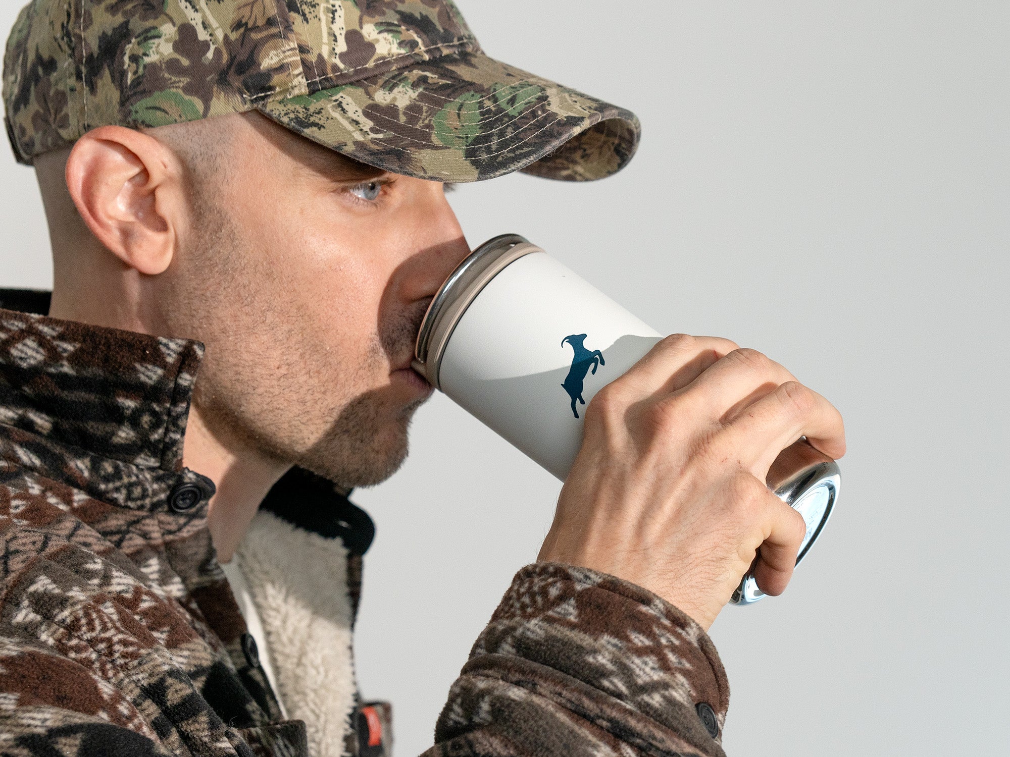 Man in camouflage gear drinking from a white tumbler with a logo.