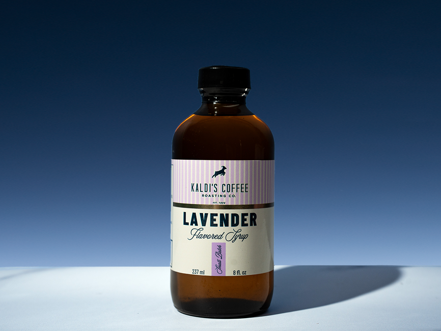 An amber glass bottle of Kaldi's Coffee Roasting Co. Lavender Flavored Syrup with a black cap, set against a dark blue gradient background with a white surface.