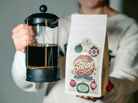 Person holding a French press with coffee and a 'Tis the Season Mocha Java' coffee bag.