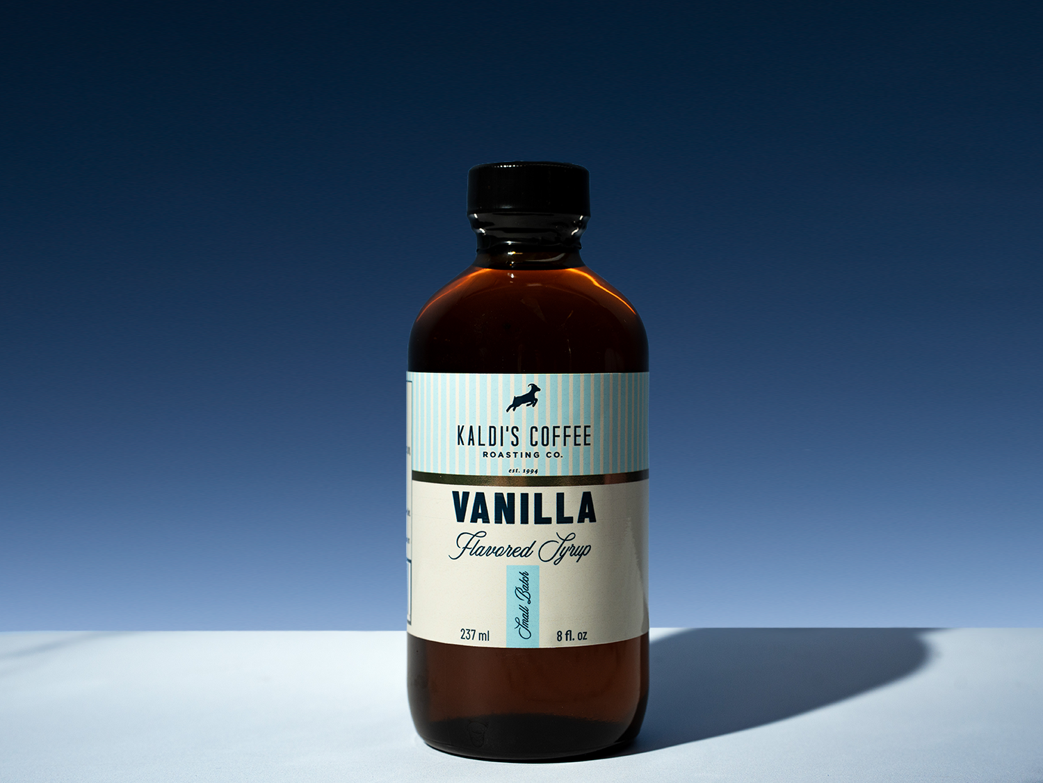 A bottle of Kaldi's Coffee Vanilla Flavored Syrup on a light surface against a dark blue gradient background. The bottle is amber glass with a black cap and a cream label with blue and black text.