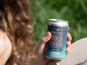 Close-up of a hand holding a chilled can of Kaldi's Coffee Cold Brew, Classic Black, with a blurred background of green grass. The can features pink "COLD BREW" lettering.