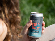 Close-up of a hand holding a chilled can of Kaldi's Coffee Cold Brew, Classic Black, with a blurred background of green grass. The can features pink "COLD BREW" lettering.