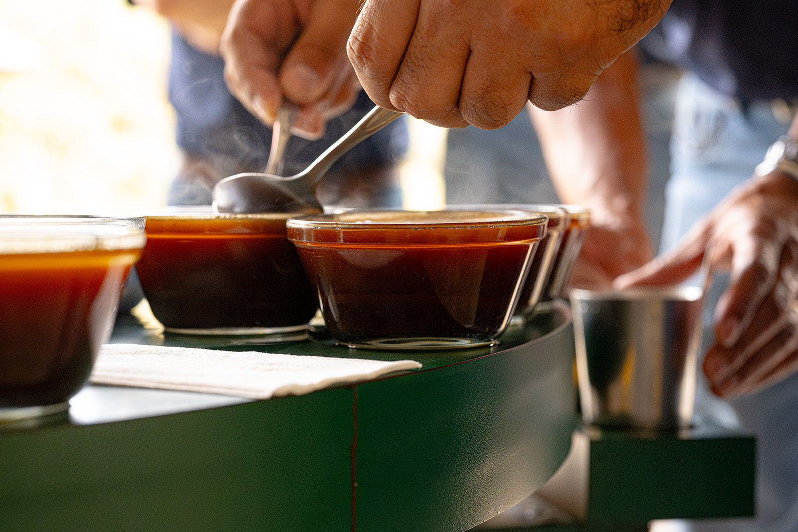 Hands hold spoons, scooping the top of a brewed cupping sample clear of grounds on a circular table.