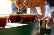 Hands hold spoons, scooping the top of a brewed cupping sample clear of grounds on a circular table.