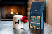 A Kaldi's Coffee diner mug with a rose next to a 12oz bag of Highlander Grogg