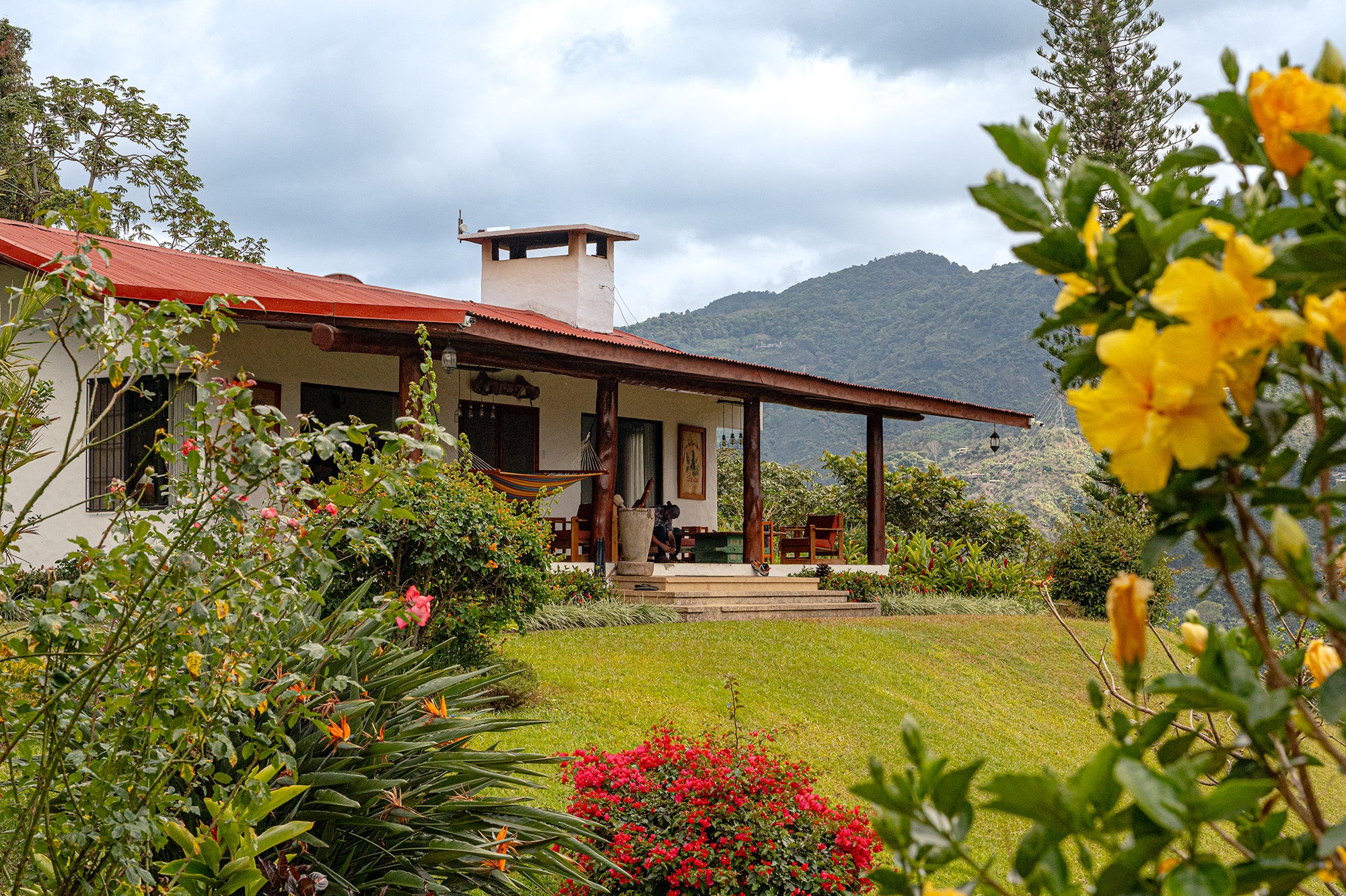 A rustic single story building with a red roof sits atop a mountain, surrounded by tropical flowers, with a higher mountain in the distance