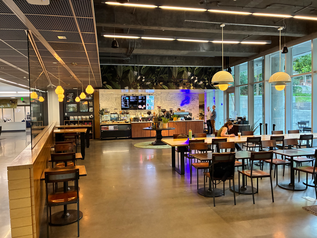 A modern cafe interior with multiple dining tables and chairs, pendant lights, a bar counter, and large windows on the right side.
