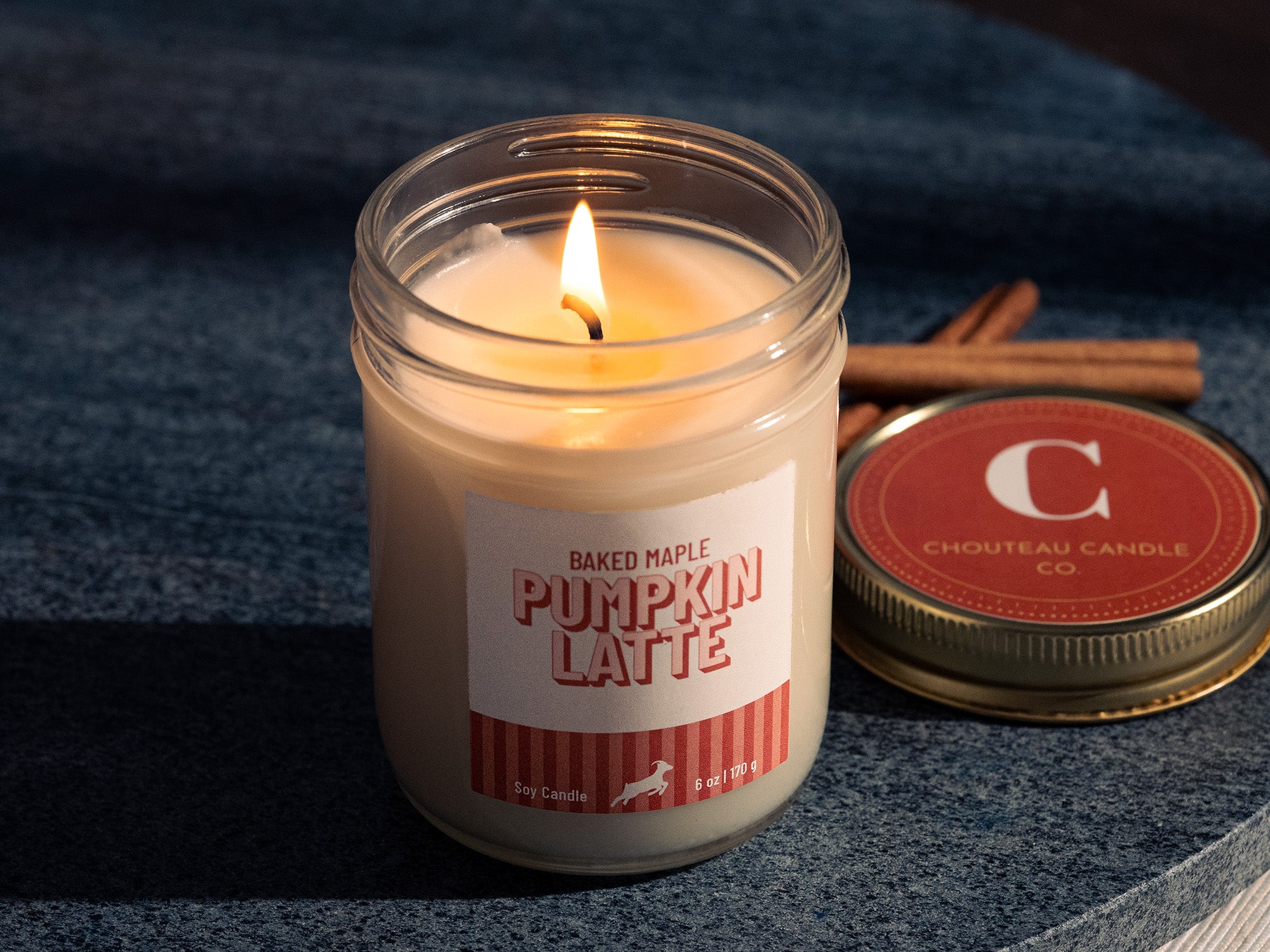 Baked Maple Pumpkin Latte candle in a glass jar with a label, surrounded by cinnamon sticks on a dark fabric background.