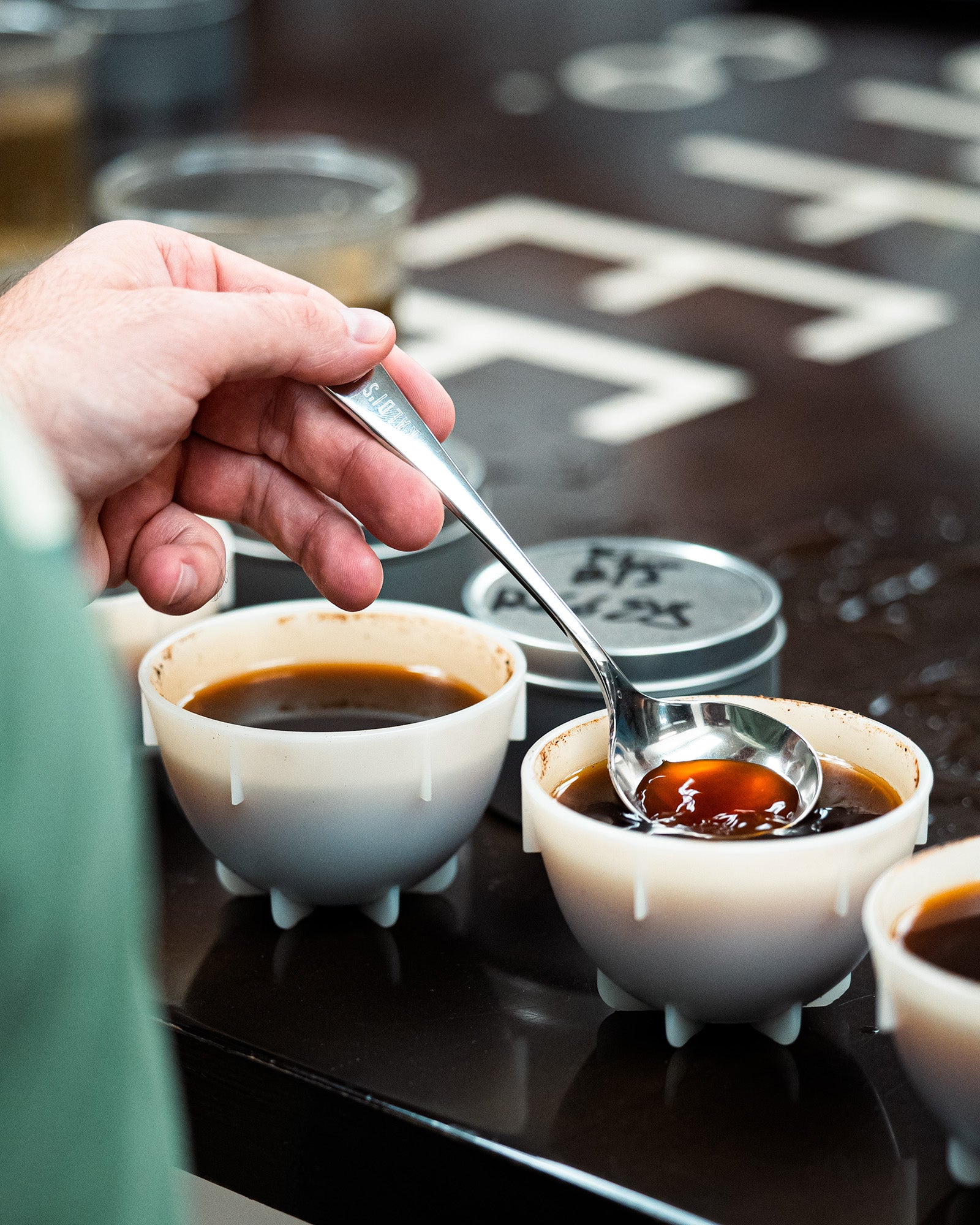 Hand holding a spoon over small bowls of coffee on a dark surface