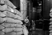 The interior of a warehouse, in black and white, showing large metal silos surrouned by 5 foot tall stacked burlap sacks of coffee. In the center, a man drags a bag of coffee from a large scale toward one stack.